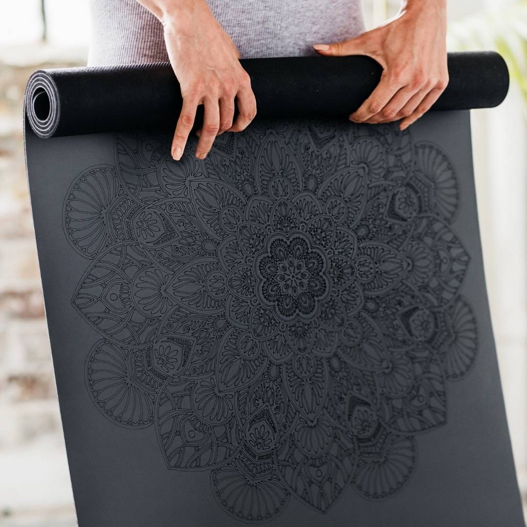 person holding a dark grey yoga mat with a Mandala pattern engraved on it